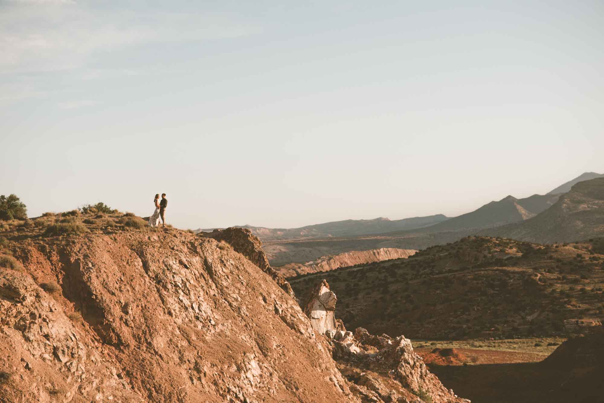 A couple stands at the edge of a high, rocky desert bluff, with a canyon and mountain ridges stretching out behind them.
