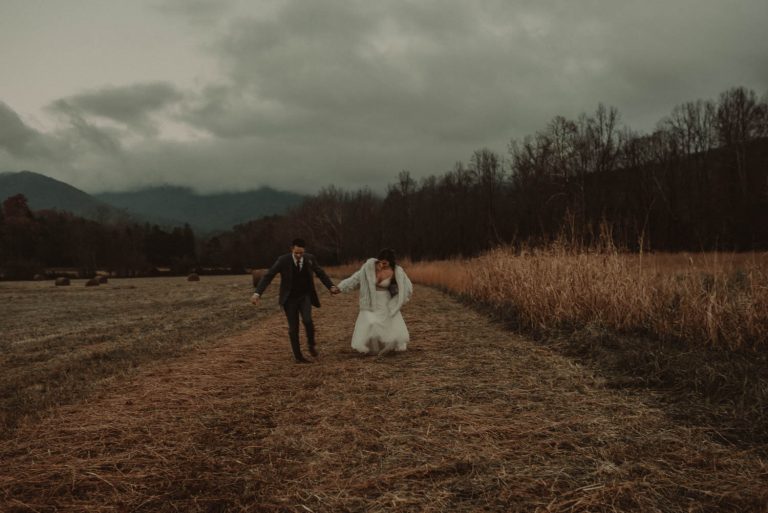 A bride in a white dress and a groom in a dark suit run along a mown path through a field toward mountains under heavy gray clouds.