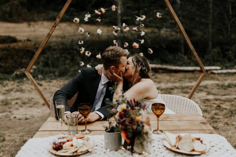 A couple kisses at an outdoor table set with flowers and glasses, paper flowers hanging above them