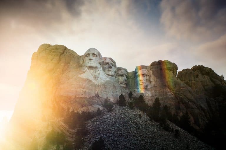 Mount Rushmore's carved presidents glow in a burst of sunlight with a faint rainbow arcing across the rock face.