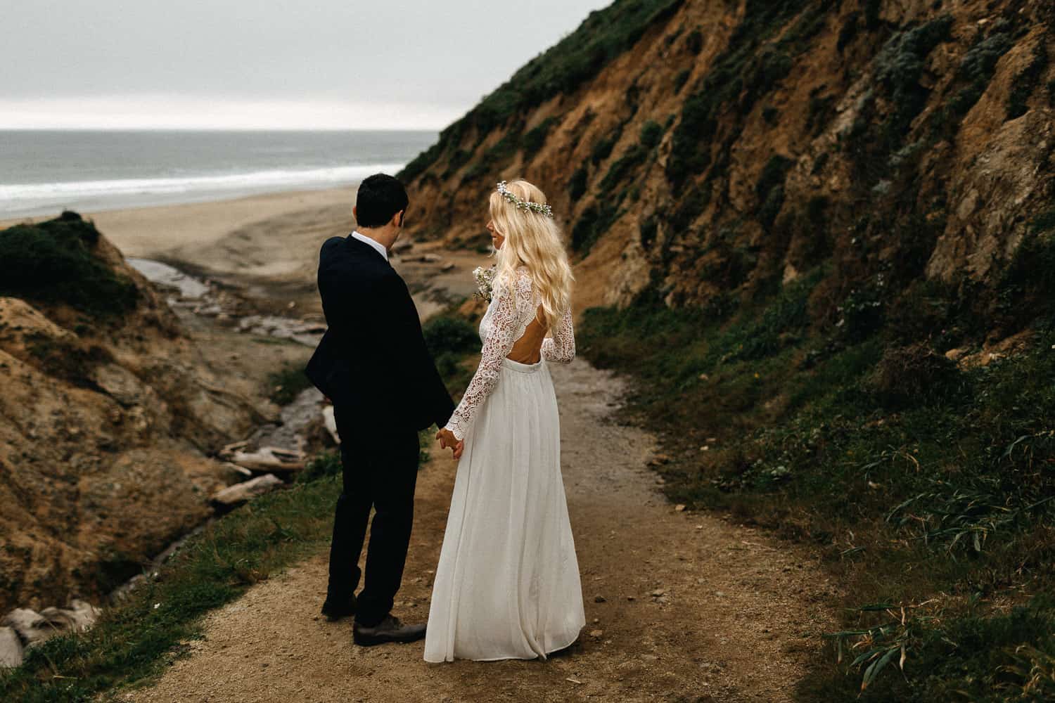 A bride in a lace-backed gown and a groom in a dark suit stand on a dirt trail above the beach between grassy cliffs at Point Reyes.