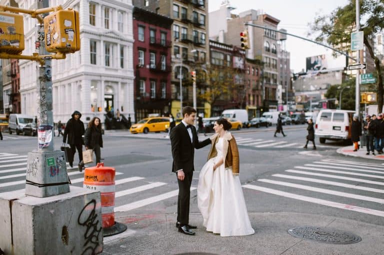 A bride in a white ball gown and brown jacket and a groom in a black tuxedo stand on a Manhattan street corner, with crosswalks, yellow taxis and brick buildings behind them.