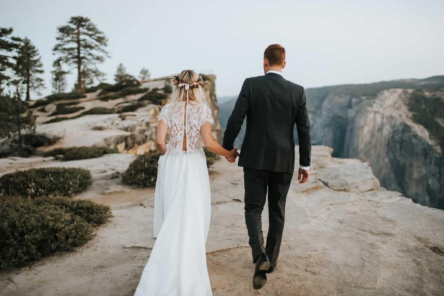 A bride with a flower crown and a groom in a dark suit walk hand in hand across the granite toward the cliffs at Taft Point in Yosemite.