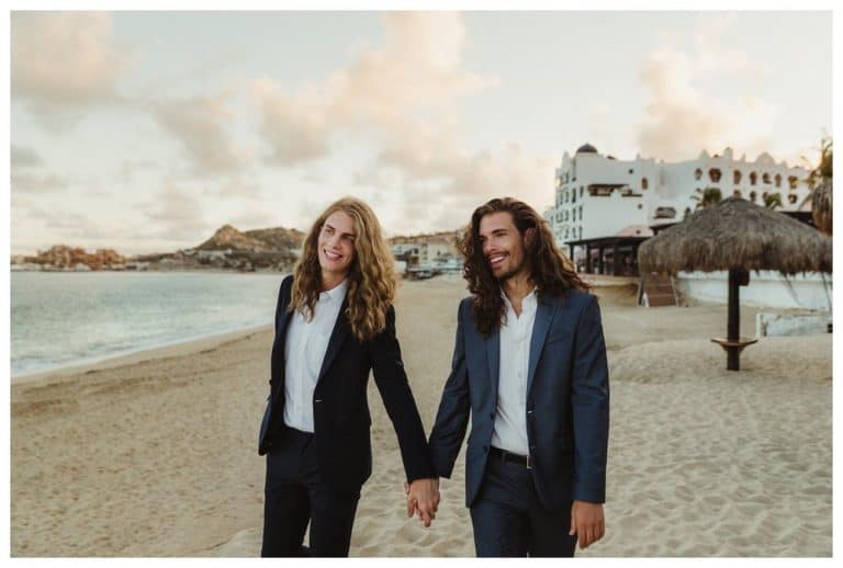 Two men in navy suits walk hand in hand along the beach in Cabo San Lucas, white buildings behind them