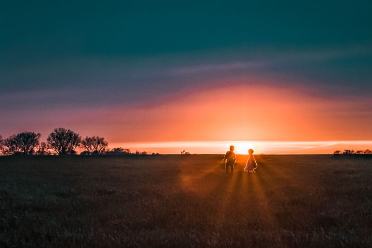 Two small figures stand in a wide, flat field, silhouetted against a deep orange sunset under a teal sky.