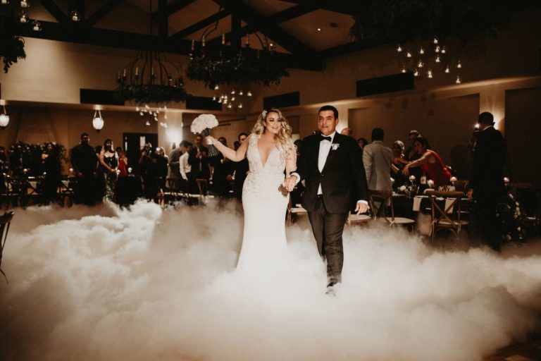 A couple walks through low fog on the dance floor of a candlelit ballroom as guests watch.