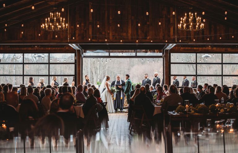 Guests at round tables watch a ceremony in front of the open doors of a barn strung with lights