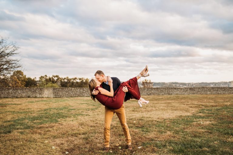 A man in mustard-colored trousers dips a woman in a burgundy dress for a kiss on a grassy field in front of a dry-stone wall and autumn trees.