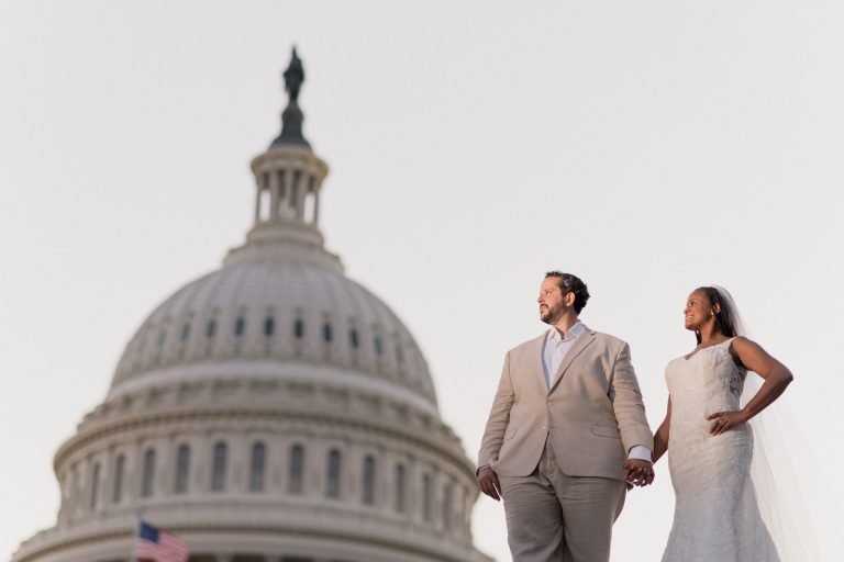 A couple in a beige suit and a white wedding dress holding hands in front of the U.S. Capitol dome
