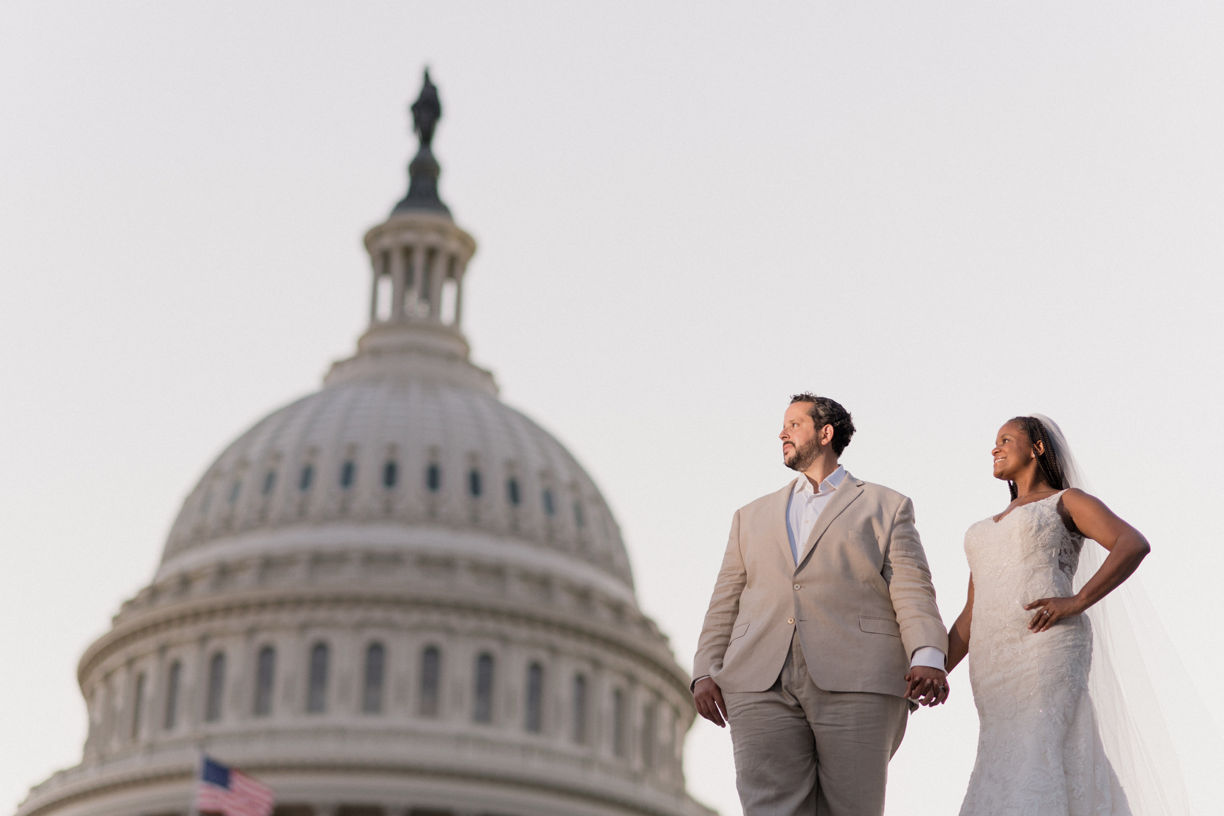 A couple in a beige suit and a white wedding dress holding hands in front of the U.S. Capitol dome