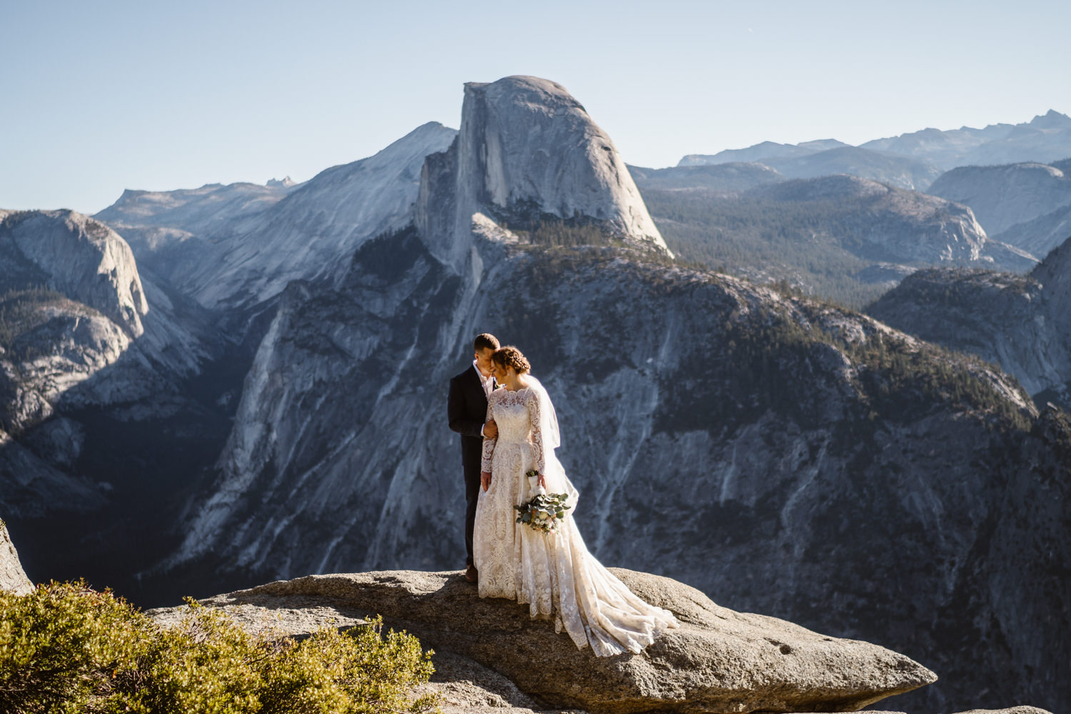 A bride in a long-sleeved lace gown and a groom in a dark suit embrace on a granite ledge at Glacier Point in Yosemite, with Half Dome rising behind them.