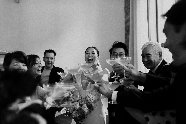 Black-and-white photo of a laughing bride raising a coupe glass with friends in a toast