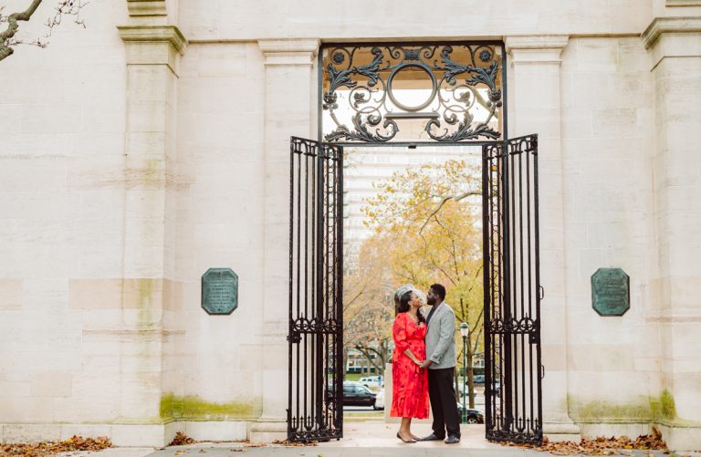 A woman in a red dress and a man in a gray suit kiss in an open wrought-iron gateway set in a pale stone wall, with autumn trees behind them.